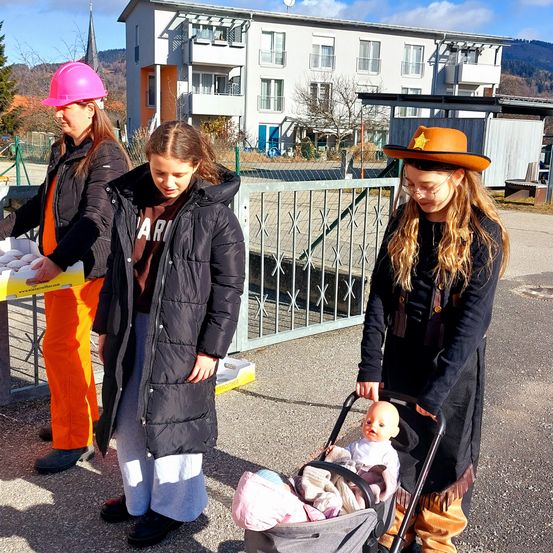 Drei Frauen stehen vor einem Gebäude. Die Frau in der Mitte schiebt einen Kinderwagen mit einem Baby und einer Puppe darin. Eine andere Frau hält eine gelbe Box. Die Frau auf der linken Seite trägt einen Helm.