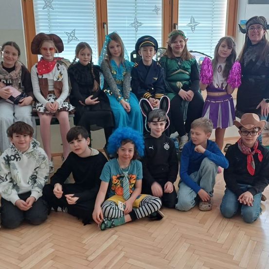 Eine Gruppe von Kindern und Erwachsenen in Kostümen posiert für ein Foto in einem Raum mit Holzböden und einem Fenster mit weißen Jalousien.