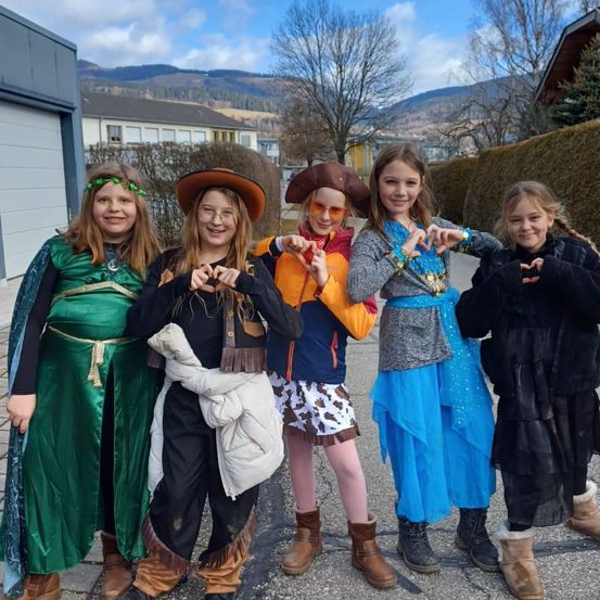 Fünf junge Mädchen in verschiedenen Kostümen posieren für ein Foto auf einer Straße, mit Bergen im Hintergrund. Jedes Mädchen macht mit ihren Händen eine Herzform.
