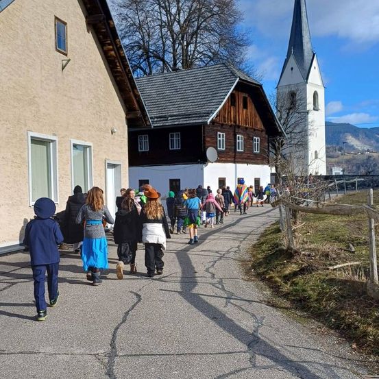 Eine Gruppe von Kindern in Kostümen geht eine Straße entlang, nahe einer Kirche und Häusern, unter klarem Himmel.