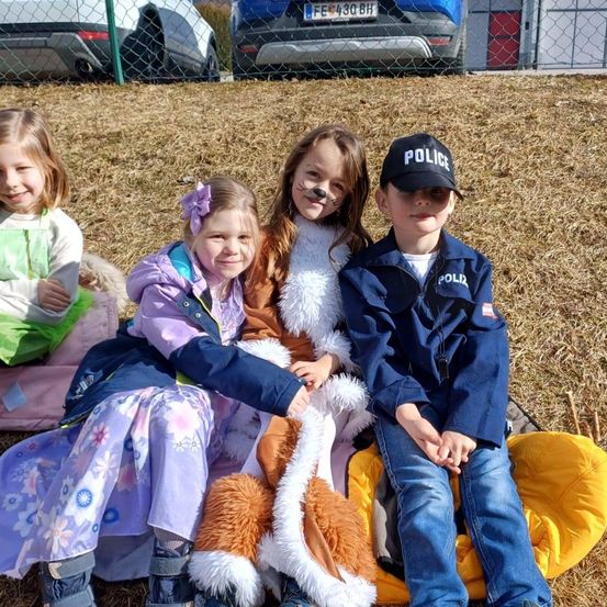 Vier Kinder, die verschiedene Kostüme tragen, sitzen auf einem Grasfeld mit getrocknetem Gras und einem Zaun im Hintergrund. Ein Kind trägt eine Polizeiuniform, während ein anderes ein pelziges Kostüm trägt. Autos sind hinter dem Zaun geparkt.