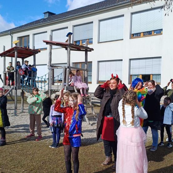 Eine Gruppe von Kindern und Erwachsenen, einige in Kostümen, versammelt sich vor einem Gebäude mit vielen Fenstern und einem Spielplatz.