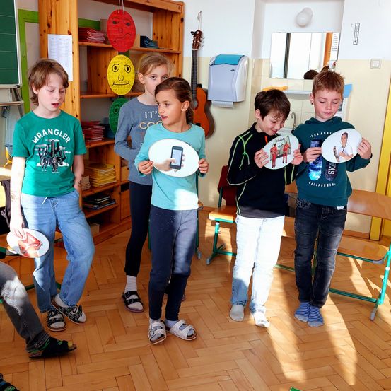 Fünf Kinder stehen in einem Klassenzimmer. Sie halten weiße Ausschnitte mit Bildern von Menschen. Der Junge auf der linken Seite trägt ein grünes T-Shirt.
