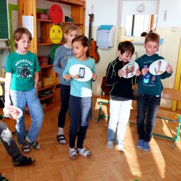 Fünf Kinder stehen in einem Klassenzimmer. Sie halten weiße Ausschnitte mit Bildern von Menschen. Der Junge auf der linken Seite trägt ein grünes T-Shirt.