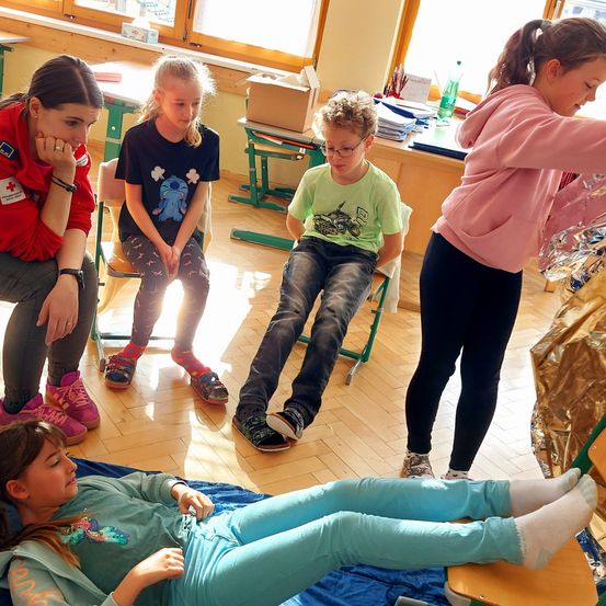 Eine Gruppe von Kindern sitzt auf Stühlen in einem Klassenzimmer. Ein Mädchen liegt auf einer blauen Matte auf dem Boden. Eine andere Mädchen steht und hält eine silberne Tasche. Ein Junge mit Brille sitzt auf einem Stuhl. Ein Mädchen schaut auf etwas auf dem Schreibtisch.