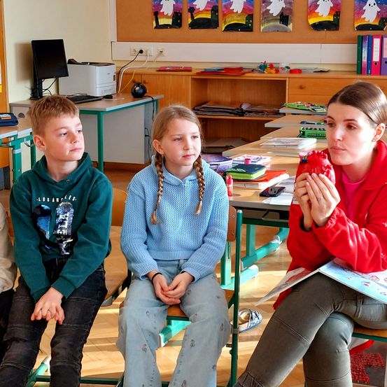 In einem Klassenzimmer liest eine Frau zwei Kindern auf Stühlen ein Buch vor. Der Raum hat Holztische, Schränke und verschiedene Gegenstände auf den Regalen. Links steht ein Drucker, und die Wand ist mit farbenfrohen Gemälden dekoriert.