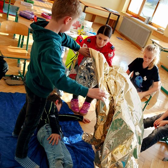 Eine Gruppe von Kindern und eine erwachsene Frau üben in einem Klassenzimmer Erste Hilfe mit einer goldenen Notfalldecke.
