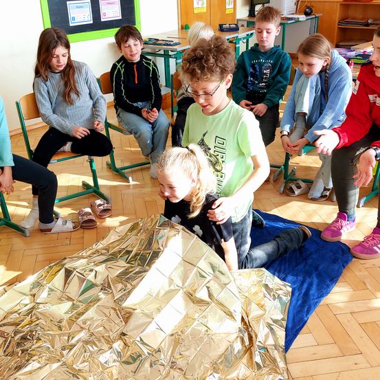 Eine Gruppe von Kindern sitzt im Kreis auf dem Boden, einige beobachten eine goldene Foliendecke auf dem Boden, während andere Brillen und Turnschuhe tragen.