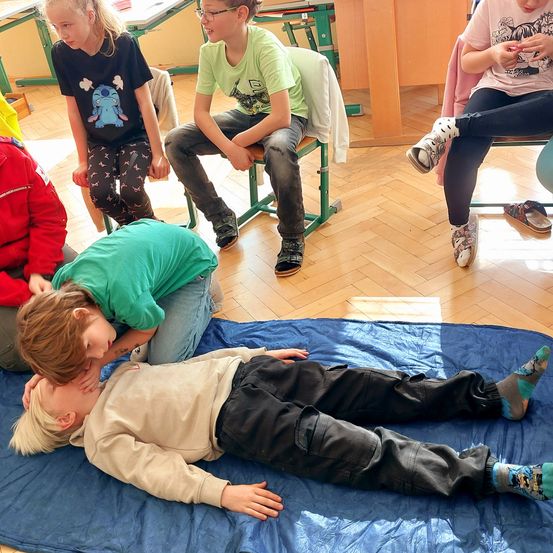In einem Klassenzimmer beobachten eine Gruppe von Kindern einen Jungen, der auf einer blauen Plane liegt. Ein anderes Kind beugt sich über ihn, während andere in Stühlen sitzen. Der Raum hat Holzböden und Schreibtische.