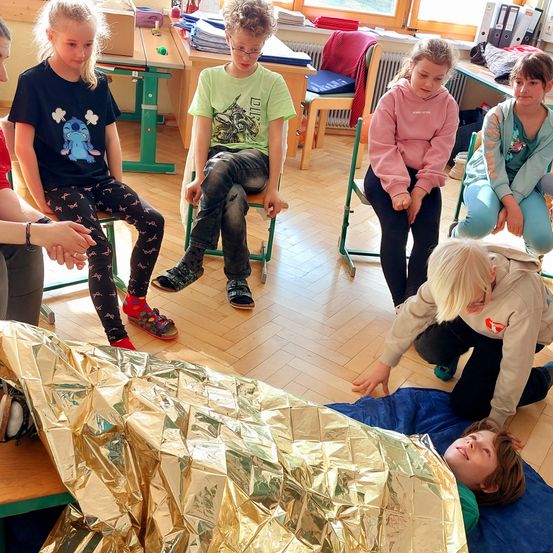 Eine Gruppe von Kindern sitzt in einem Kreis um einen Jungen, der auf einer blauen Decke liegt, die mit einer goldenen Folie bedeckt ist. Sie scheinen an einer Aktivität teilzunehmen.