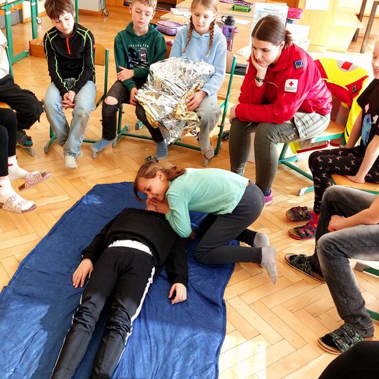 Mehrere Kinder sitzen um eine zentrale Figur herum, die auf einem blauen Teppich liegt, möglicherweise in einem Klassenzimmer. Die liegende Person scheint von einem Mädchen Aufmerksamkeit zu erhalten. Eine andere Mädchen sitzt in der Nähe mit einer reflektierenden Decke. Eine Frau in einer roten Jacke beobachtet die Szene.