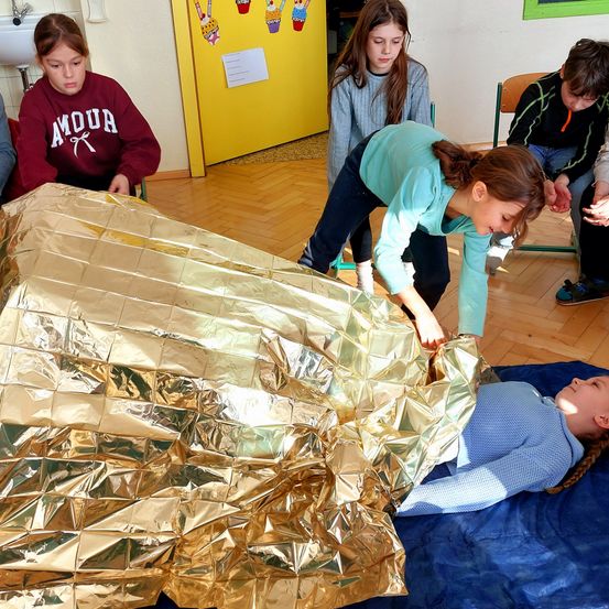 Eine Gruppe von Kindern ist in einem Raum, wobei eines auf einem blauen Laken liegt, das mit Goldfolie bedeckt ist. Sie beobachten und interagieren mit der Einrichtung.