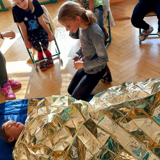 Eine Gruppe von Kindern ist drinnen, ein Kind liegt auf einem blauen Tuch, das mit einer goldenen Notfalldecke bedeckt ist. Andere Kinder sitzen auf Stühlen und beobachten.