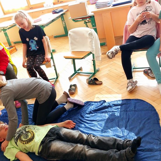 In einem Klassenzimmer sitzen mehrere Kinder, einige ohne Schuhe, ein Kind liegt auf einer blauen Decke und ein anderes beugt sich, um zu helfen.