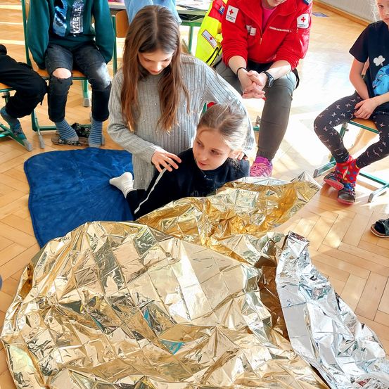 Mehrere Kinder sind drinnen; ein Kind übt, ein anderes in eine goldene Foliendecke zu wickeln. Das Kind sitzt, und eine Frau steht neben ihr.