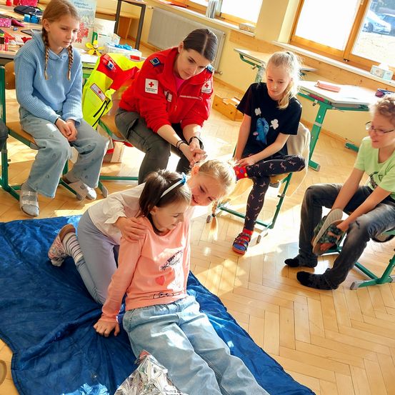 Eine Frau in einer roten Jacke hilft einem kleinen Mädchen, das auf einer blauen Decke in einem Klassenzimmer liegt. In der Nähe sitzen Kinder auf Stühlen, eines der Mädchen trägt eine Brille. Das Klassenzimmer hat gelbe Wände und Holzböden.