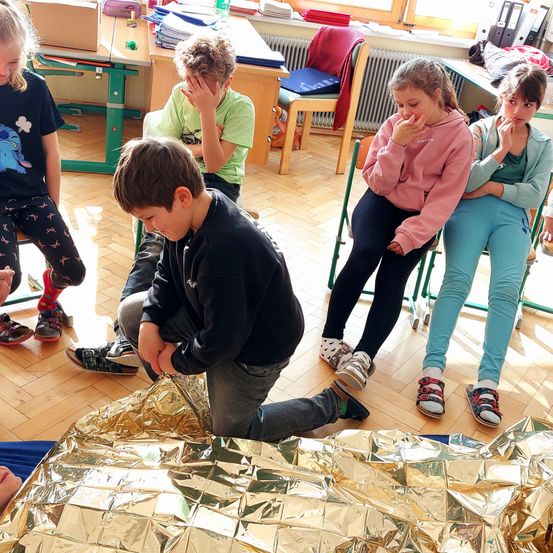 Eine Gruppe von Kindern sitzt auf dem Boden, zwei kniend, eines hält Folie. Drei Mädchen sitzen auf Stühlen, eines in Pink, ein anderes in Blau. Ein Tisch und ein Stuhl stehen dahinter.