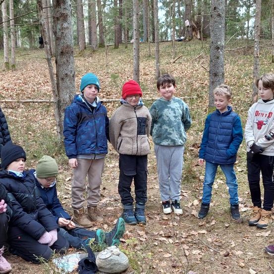 Eine Gruppe von Kindern, einige sitzend und andere stehend, posiert für ein Foto in einem bewaldeten Gebiet, umgeben von gefallenen Blättern.