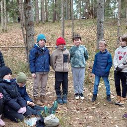 Eine Gruppe von Kindern, einige sitzend und andere stehend, posiert für ein Foto in einem bewaldeten Gebiet, umgeben von gefallenen Blättern.