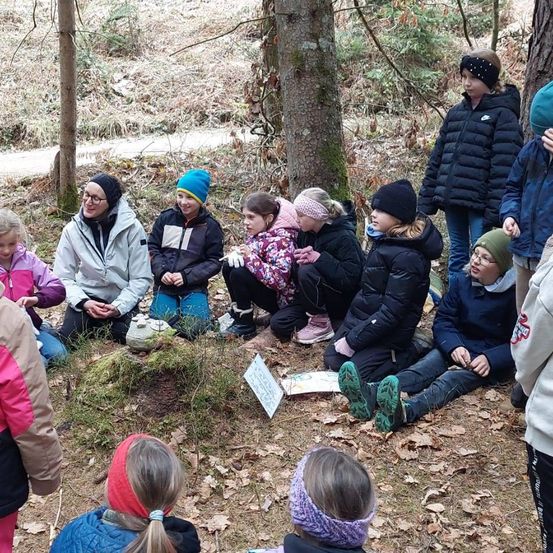 Eine Gruppe von Kindern und Erwachsenen in Winterkleidung sitzt in einem Kreis im Wald und hört einer Frau zu, die ein Schild hält.