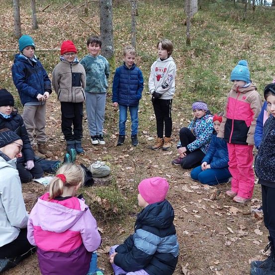 Eine Gruppe von Kindern in Winterkleidung, einige stehend und andere sitzend, in einem bewaldeten Bereich. Sie befinden sich wahrscheinlich auf einer Schulausflugstour.