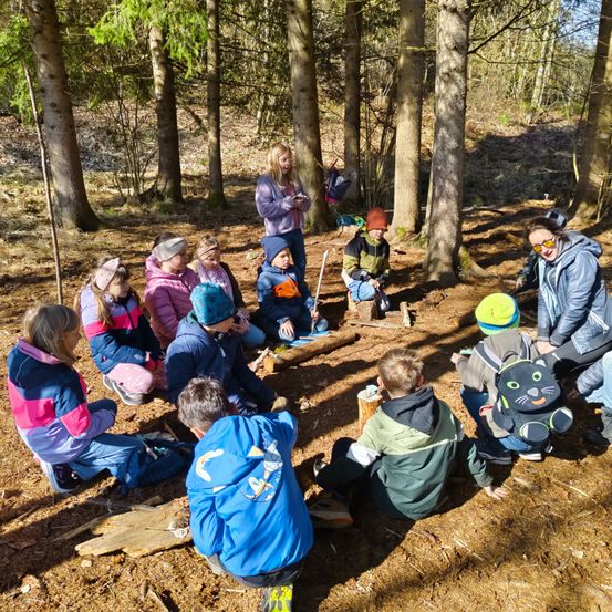 Eine Gruppe von Kindern und Erwachsenen sitzt im Kreis in einer Waldlichtung, trägt Winterkleidung und teilweise Mützen. Eine Frau mit Brille steht in der Nähe.