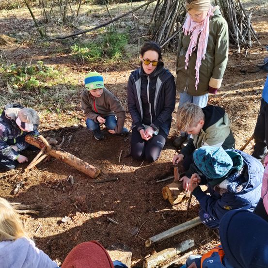 Eine Gruppe von Kindern und einer erwachsenen Frau befindet sich im Wald und arbeitet mit Holzstämmen und Werkzeugen. Sie tragen Winterkleidung.