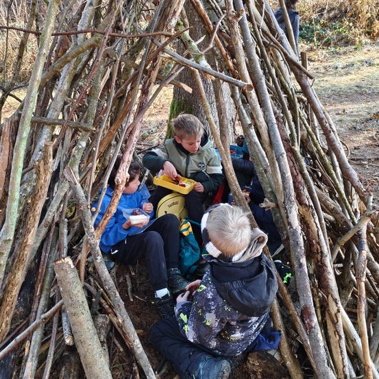 Drei Kinder sitzen in einem aus Stöcken gebauten Unterschlupf. Ein Kind isst von einem gelben Tablett, ein anderes hält einen Behälter und das dritte beobachtet. Sie tragen alle Jacken.