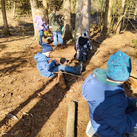 Kinder in Winterkleidung sind in einem Wald, einige sitzen und andere stehen. Ein Kind hält einen Stock, ein anderes ist in der Nähe eines Baumstamms. Der Boden ist mit Schmutz und Blättern bedeckt.