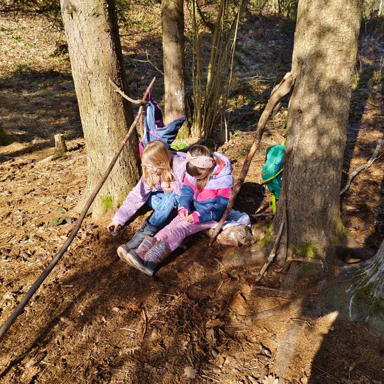 Zwei Kinder sitzen auf dem Boden in einem Wald in der Nähe eines Baumes und lächeln, während sie etwas in den Händen halten.