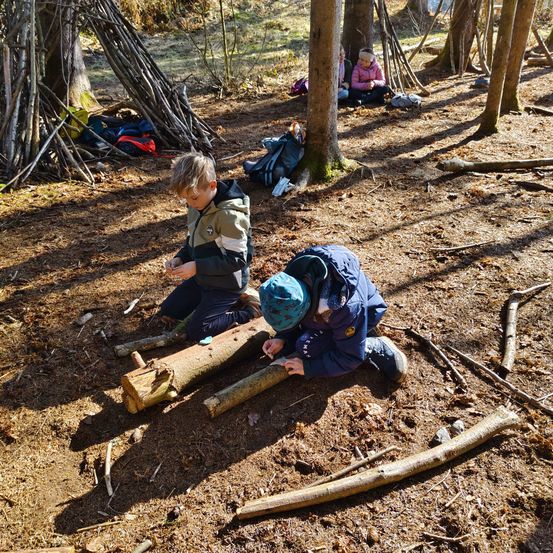 Zwei Kinder sitzen auf Baumstämmen in einem Wald, wahrscheinlich bei einer Bastel- oder Outdoor-Aktivität. In der Nähe sitzt eine Person auf dem Boden. Der Waldboden ist mit Ästen und Blättern bedeckt.