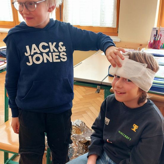 Zwei Jungen in einem Klassenzimmer, einer mit einem Verband am Kopf, der andere hält seinen Kopf.