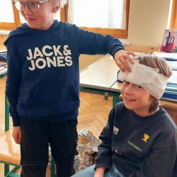 Zwei Jungen in einem Klassenzimmer, einer mit einem Verband am Kopf, der andere hält seinen Kopf.