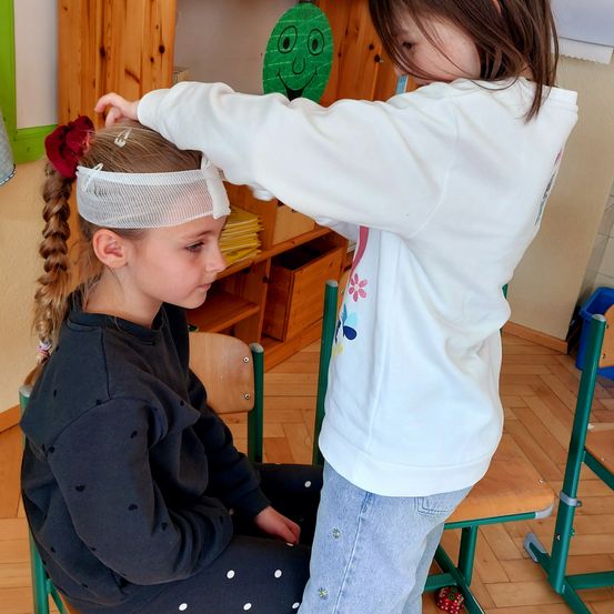 Ein junges Mädchen sitzt auf einem Stuhl mit einem Verband auf dem Kopf. Ein anderes Mädchen steht hinter ihr und legt ihr einen Verband an. Hinter ihnen sind ein Holzregal und ein Stuhl zu sehen.