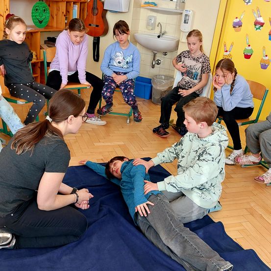 Eine Gruppe von Kindern und einer Frau sind in einem Klassenzimmer. Sie sitzen auf Stühlen um einen Jungen, der auf einer blauen Matte liegt. Eine Frau hält die Hand des Jungen. Eine Gitarre hängt an der Wand und ein Waschbecken ist an der Wand.