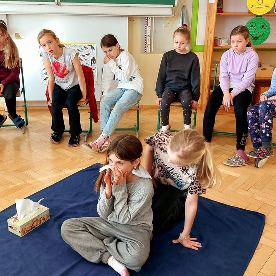 Eine Gruppe von Mädchen sitzt in einem Klassenzimmer auf Stühlen, manche mit gesenktem Kopf. Ein Mädchen schnäuzt sich, ein anderes beugt sich über sie. Eine Schachtel Taschentücher liegt auf dem Boden.