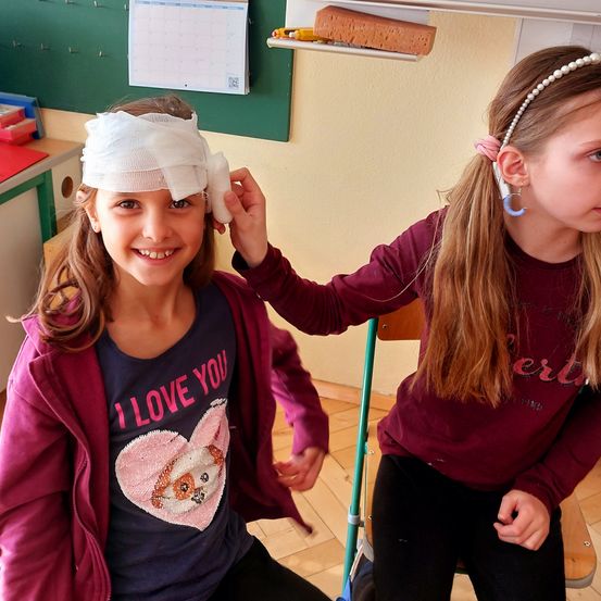 Zwei junge Mädchen in einem Klassenzimmer. Eines der Mädchen hat einen Verband auf dem Kopf, und das andere Mädchen hilft dabei.