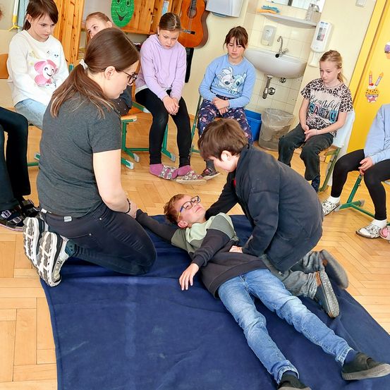 Eine Gruppe von Kindern sitzt im Kreis und beobachtet eine Frau und einen Jungen, die auf einem blauen Teppich auf dem Boden liegen, während ein Junge über ihnen steht.