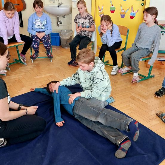 Eine Gruppe von Kindern in einem Klassenzimmer beobachtet, wie ein Junge auf einer blauen Matte liegt, unterstützt von einem anderen Kind. Eine Frau mit Brille beobachtet ebenfalls.