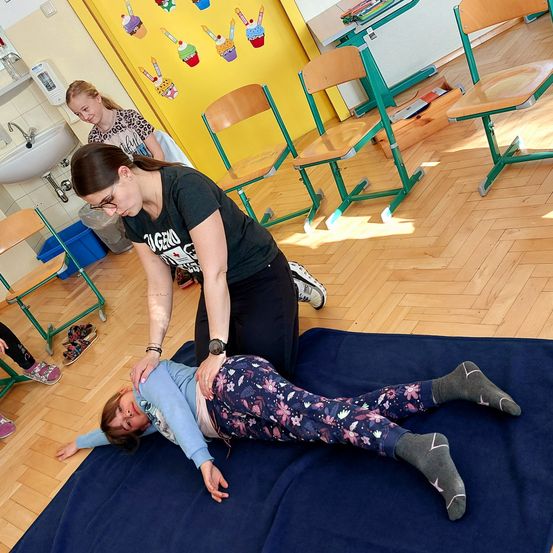 Eine Frau hilft einem Kind, das auf einem blauen Teppich auf dem Boden in einem Klassenzimmer mit Holzstühlen und einer gelben Wand mit Cupcake-Bildern liegt.