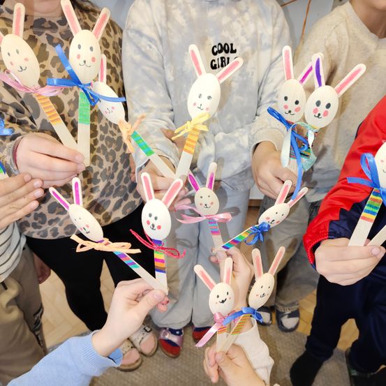 Eine Gruppe von Kindern hält Osterhasen-Themen-Holzstäbe mit bunten Bändern, einige in Tierdruckkleidung, einem 'Cool Girl' Sweatshirt und Turnschuhen.