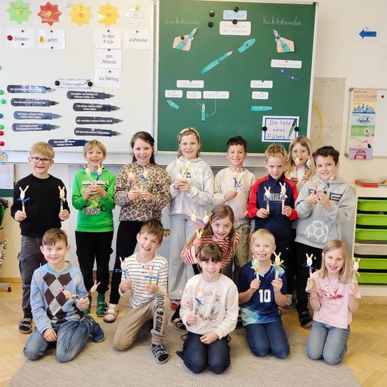Eine Gruppe von Kindern im Klassenzimmer, die selbstgemachte Gegenstände halten, posiert für ein Foto. Hinter ihnen befindet sich eine grüne Tafel mit verschiedenen Aufklebern und Wörtern.