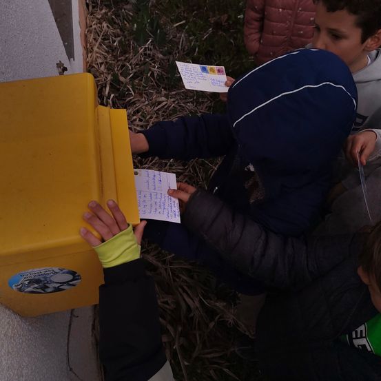 Vier Kinder legen Briefe in eine gelbe Box. Zwei der Kinder tragen Handschuhe. Ein Kind hält einen Brief in jeder Hand.