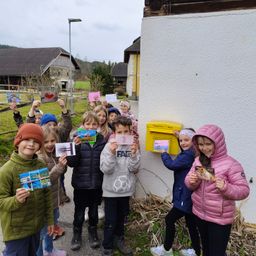 Eine Gruppe von Kindern in Winterkleidung steht vor einem Haus und hält Postkarten in der Hand. Ein gelber Briefkasten ist an der Wand zu sehen.