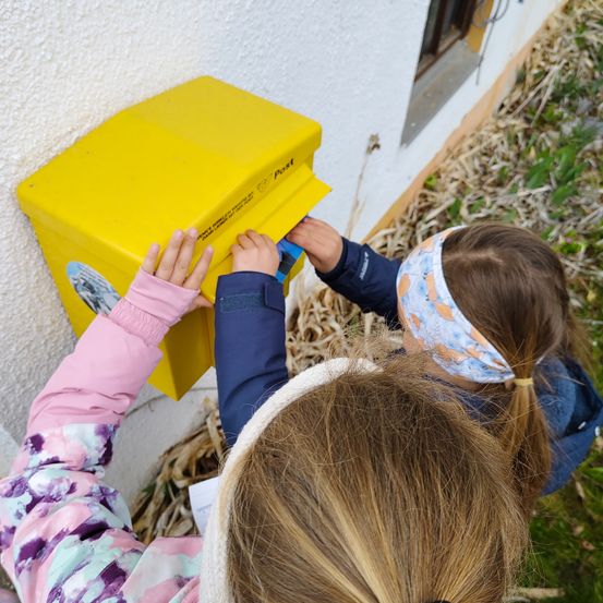 Zwei junge Mädchen stecken einen Brief in einen gelben Briefkasten an einer weißen Wand. Ein Mädchen trägt einen Haarreif.