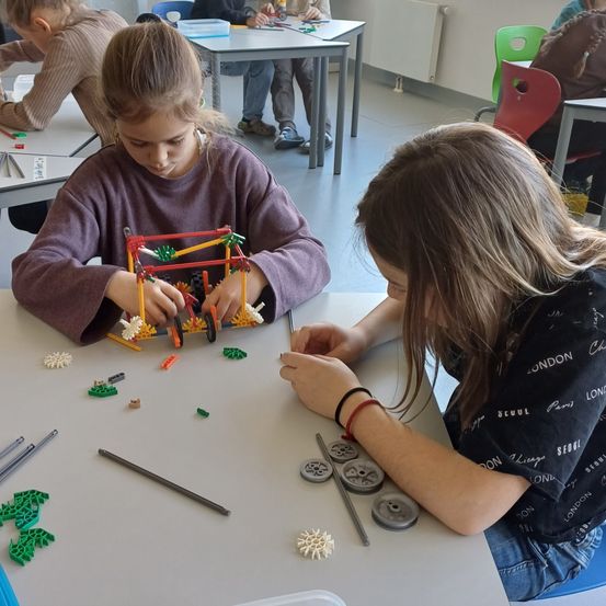 Zwei junge Mädchen arbeiten an einem LEGO-Projekt an einem Schreibtisch in einem Klassenzimmer, umgeben von anderen Schülern.