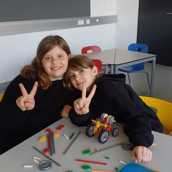 Zwei junge Mädchen sitzen an einem Tisch mit verschiedenen Lego-Steinen. Sie lächeln und machen Friedenszeichen. Der Tisch hat einen Lego-Wagen und andere Lego-Stücke. Der Raum hat Tische und Stühle.