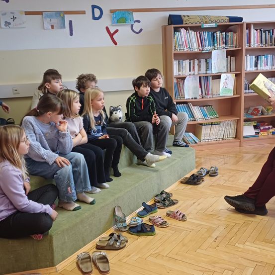 Eine Gruppe von Kindern hört in einem Klassenzimmer einer Geschichte zu. Einige sitzen auf einer Bank, andere auf dem Boden, und viele Paar Schuhe sind davor platziert.