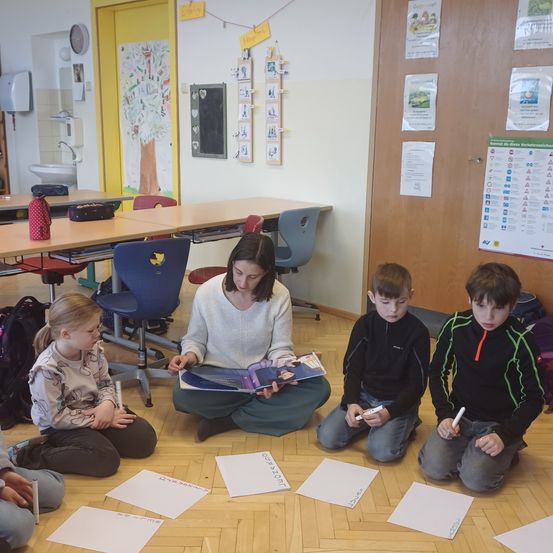 Eine Frau liest drei kleinen Kindern in einem Klassenzimmer ein Buch vor. Jedes Kind hält einen Marker und Papier. Im Hintergrund befinden sich Stühle, Schreibtische und Anschlagtafeln.