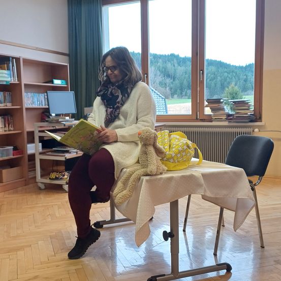 Eine Frau sitzt in einem Klassenzimmer, liest ein Buch und trägt eine Brille. Ein Teddybär ruht auf einem Tisch, und ein Stuhl steht in der Nähe. Hinter ihr zeigt ein Fenster die Aussicht auf Berge.